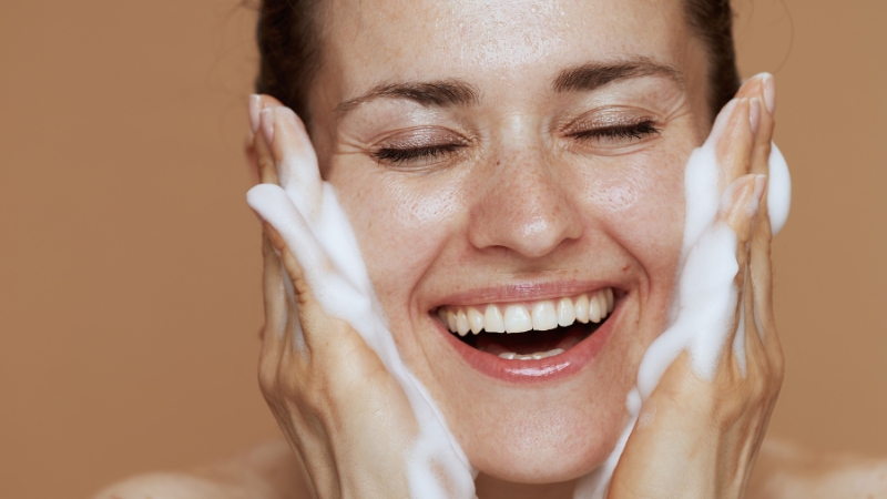 A woman smiling while applying foam from drugstore facial cleansers to her face