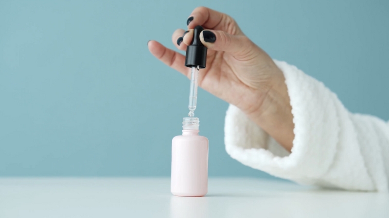 A person holds a dropper above a pink bottle while inspecting a liquid skincare product