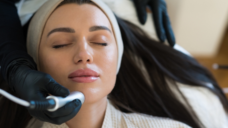 A woman relaxes with her eyes closed while a professional performs a HydraFacial treatment using a small handheld device
