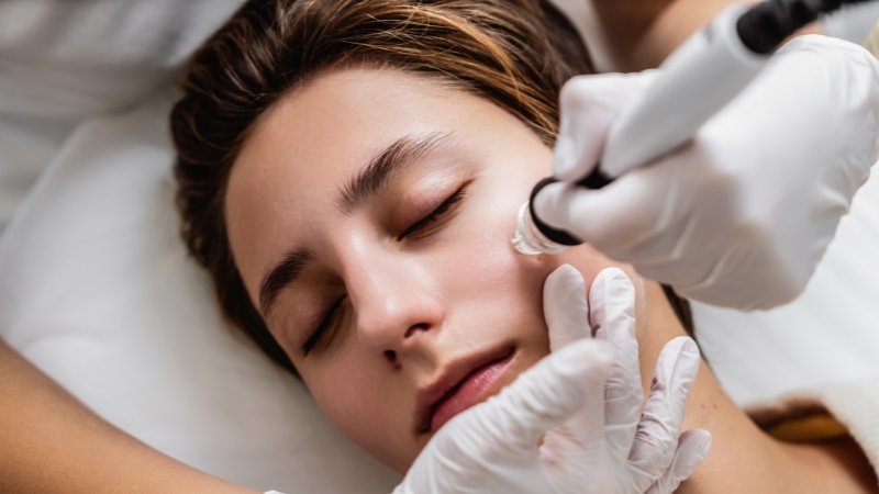A person receives a HydraFacial treatment on the cheek with a professional using a handheld device
