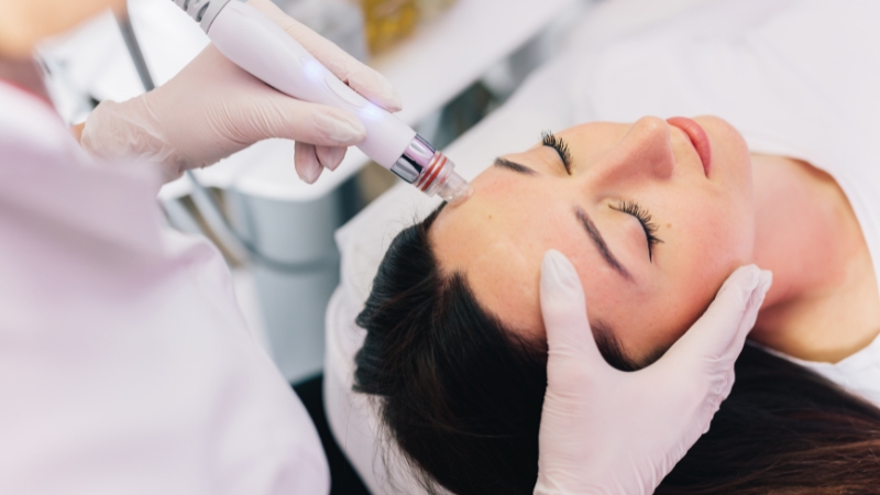 A woman receives a HydraFacial on her forehead while lying comfortably with her eyes closed