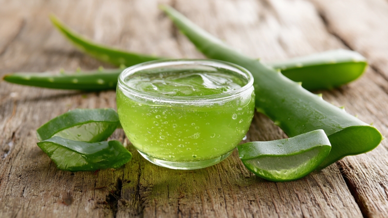 A small glass bowl of fresh green aloe vera gel placed on a rustic wooden table beside sliced aloe vera leaves