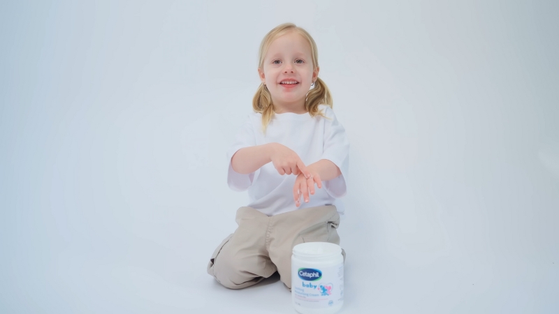 A smiling child applies moisturizer beside a jar of gentle skincare cream