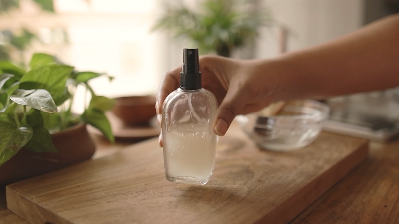 A hand holding a spray bottle filled with aloe vera gel on a wooden surface near green plants