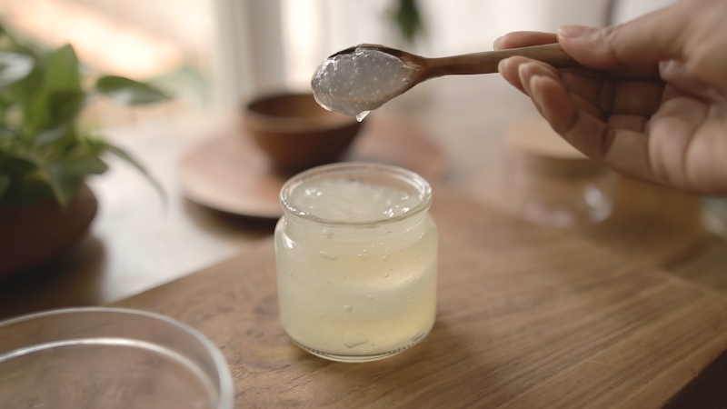 A person scooping aloe vera gel from a small glass jar with a wooden spoon on a wooden table