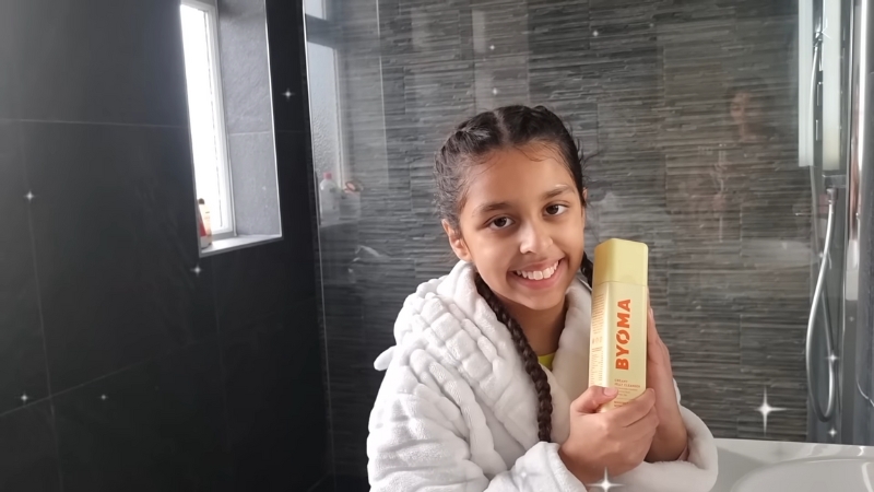 A young girl in a bathrobe holds a BYOMA skincare product